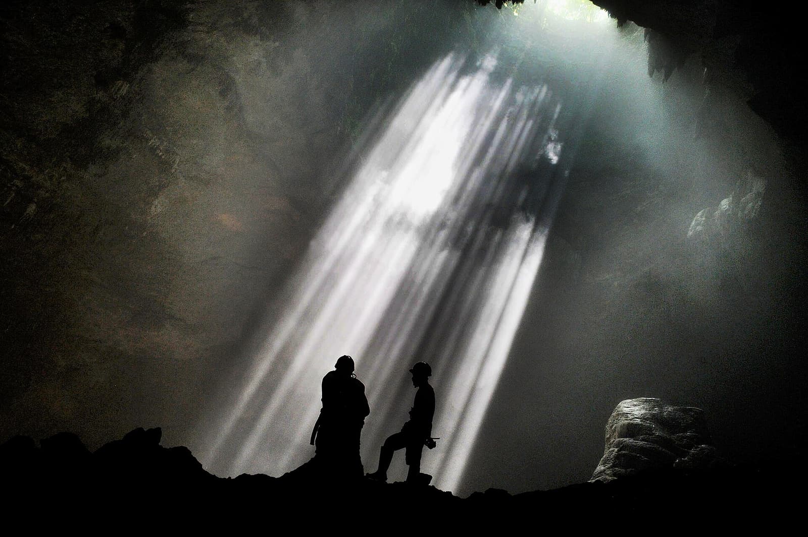 Two visitors silhouetted against the sunbeams falling into the cave at Jomblang