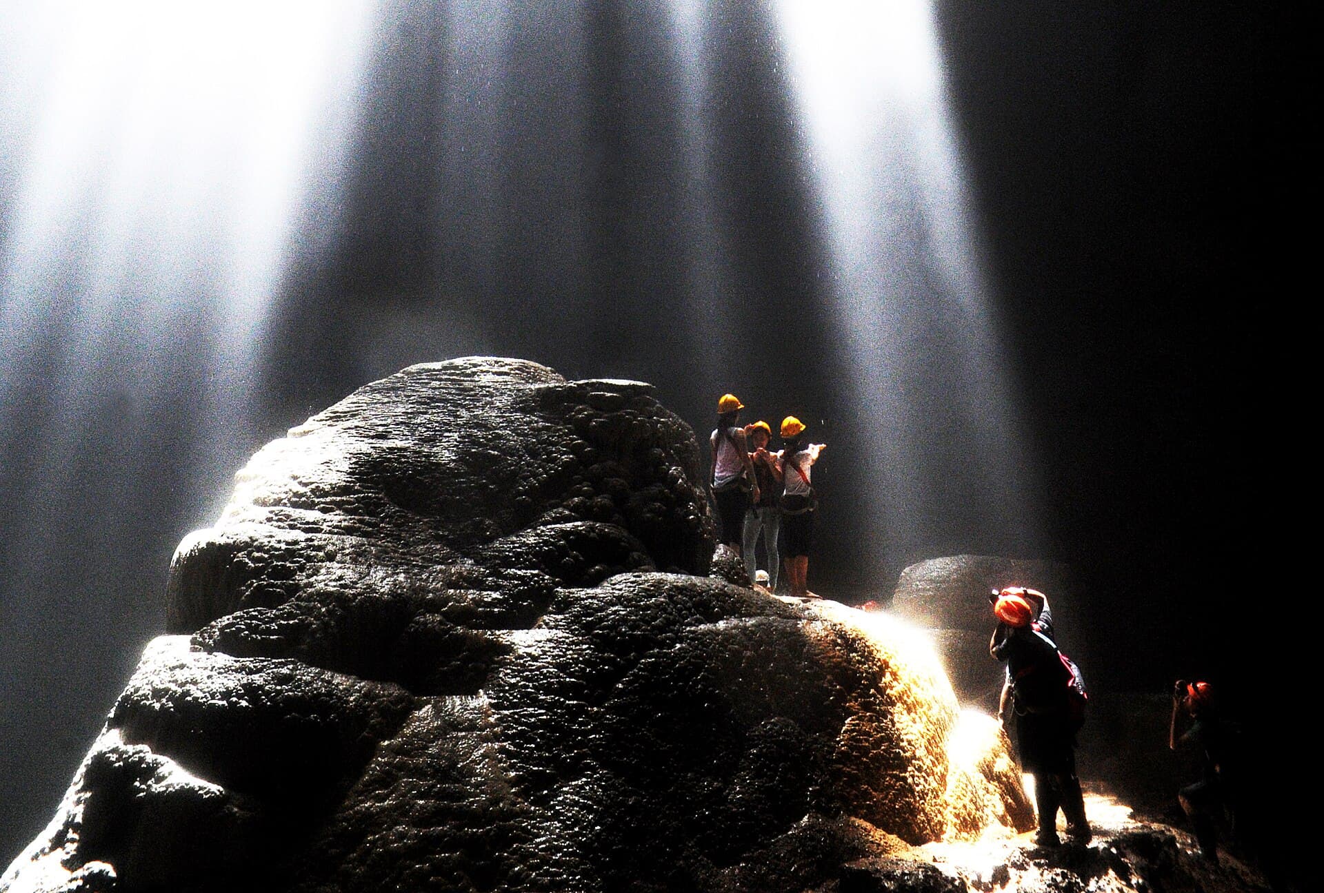 Visitors in helmets standing on the rocks beneath shafts of sunlight inside Grubug Cave at Jomblang