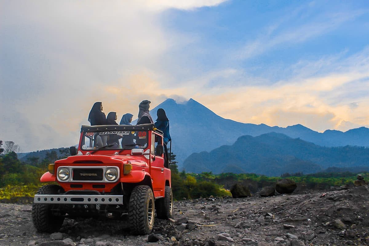 Visitors sitting on a red open-top jeep on the Merapi lava tour, with the volcano behind